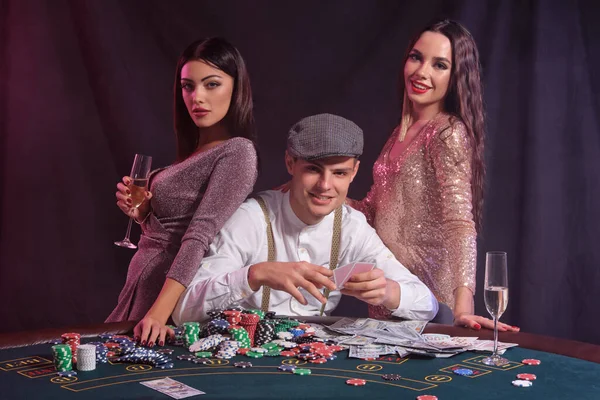 男性は チップ カードの山とテーブルに座っているカジノでポーカーをプレイしています 2人の笑顔の女性との勝利を祝う 黒の背景 カラフルなバックライト ギャンブル アルコール — ストック写真