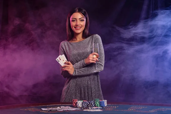 カジノでポーカーをプレイグレーのドレスでかなり乙女 シャンパンのガラスを保持 2つのエース チップ カードのスタックをテーブルに配置します 煙の背景 カラフルなバックライト — ストック写真