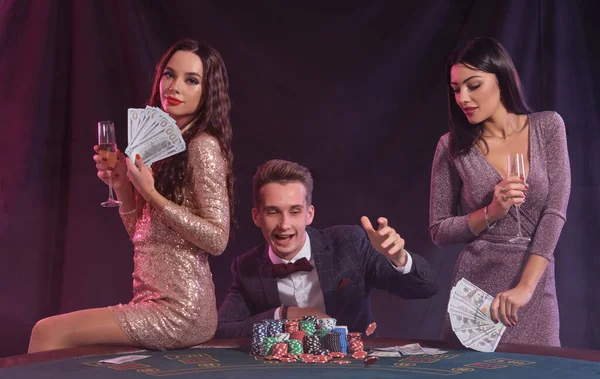 男はチップ カードの山とテーブルに座ってカジノでポーカーをプレイしています 2人の女性が現金を持って勝利を祝う 黒の背景 カラフルなバックライト ギャンブル アルコール — ストック写真