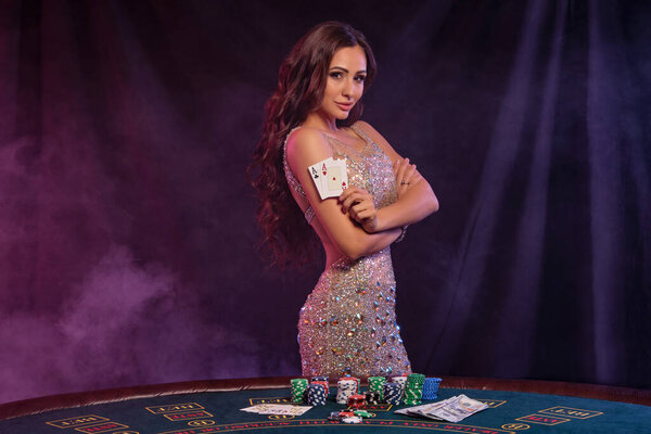 Charming female in golden dress playing poker at casino, holding two aces. Posing at table with stacks of chips, money on it. Black, smoke background, colorful backlights. Close-up.