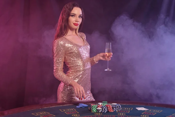 Beautiful lady in golden dress holding chip and glass of champagne, posing sideways at playing table in casino. Black, smoke background, colorful backlights. Gambling entertainment, poker. Close-up.