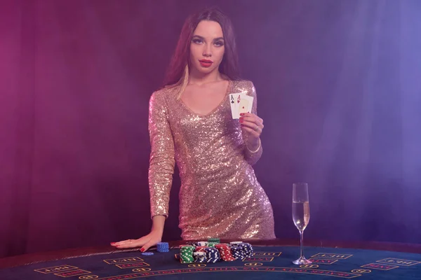Gorgeous lady in golden dress is showing two cards while posing at playing table in casino. Black, smoke background with colorful backlights. Gambling entertainment, poker, champagne. Close-up.