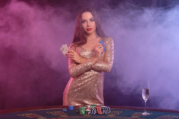 Женщина в золотом платье показывает фишки и карты, позируя за игровым столом в казино. Черный, дымовой фон. Азартные игры, покер, шампанское. Крупный план . — стоковое фото