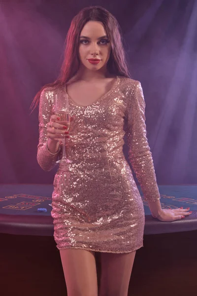 Egy arany ruhás nő kártyázik, pezsgőt tart, miközben a kaszinóban pózol. Fekete, füst háttér. Szerencsejáték, póker. Közelkép. — Stock Fotó