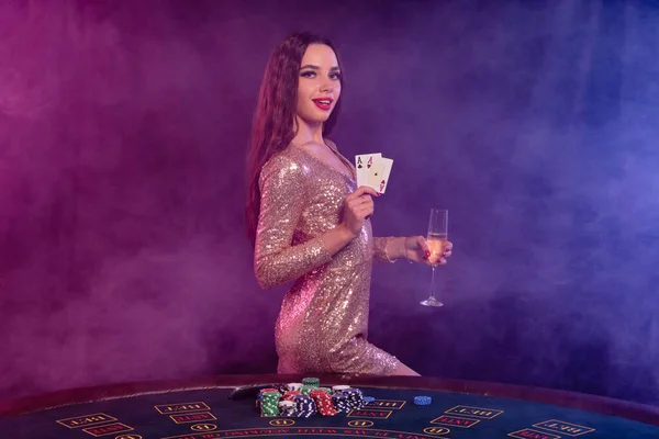 Chica jugando al póquer en el casino, sosteniendo una copa de champán, dos ases. Posando en la mesa con papas fritas, dinero encima. Negro, fondo de humo. Primer plano . —  Fotos de Stock