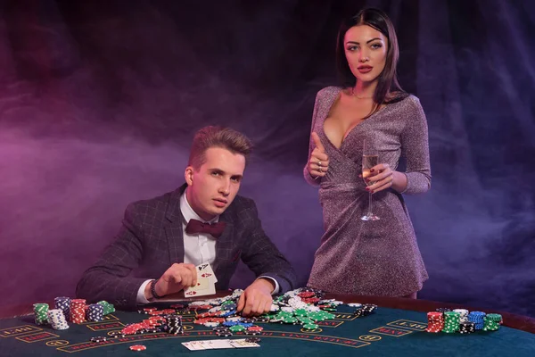 Hombre jugando al póquer en el casino sentado en la mesa con montones de fichas, dinero, champán, cartas. Celebrando la victoria con la mujer. Fondo negro. Primer plano . — Foto de Stock