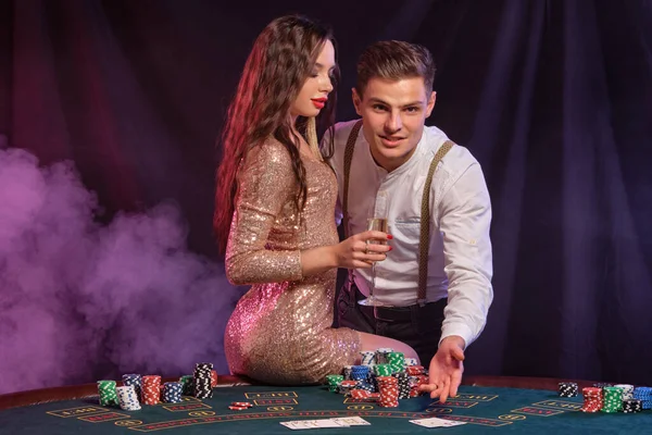 Muž a žena hrají poker v kasinu, slaví vítězství u stolu s hromadami žetonů, karet, šampaňského. Černá, kouřové pozadí. Detailní záběr. — Stock fotografie