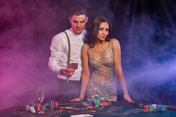Man and woman playing poker at casino, celebrating win at table with stacks of chips, cards, champagne. Black, smoke background. Close-up.