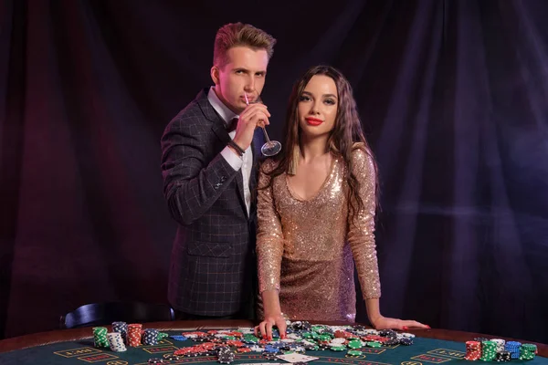 Muž a žena hrají poker v kasinu, slaví vítězství u stolu s hromadami žetonů, peněz, karet, šampaňského. Černá, kouřové pozadí. Detailní záběr. — Stock fotografie