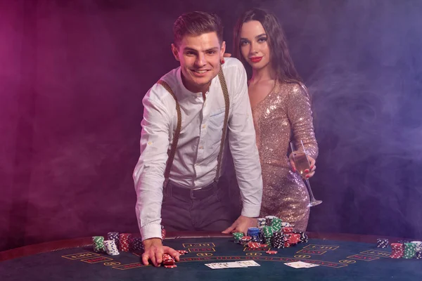 Man and woman playing poker at casino, celebrating win at table with stacks of chips, cards, champagne. Black, smoke background. Close-up. — Stock Photo, Image