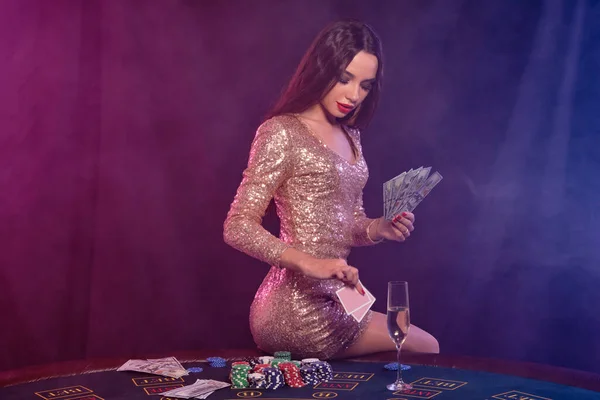 Mädchen spielen Poker im Casino, halten Geld, Karten. Mit Chips, Champagner und Bargeld am Tisch sitzen. Schwarzer, rauchiger Hintergrund. Nahaufnahme. — Stockfoto