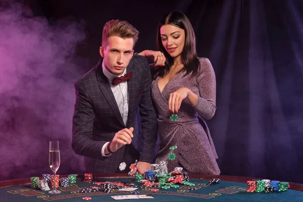 Muž a žena hrají poker v kasinu, slaví vítězství u stolu s hromadami žetonů, peněz, karet, šampaňského. Černá, kouřové pozadí. Detailní záběr. — Stock fotografie