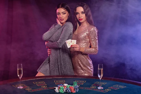 Dvě dívky hrají poker v kasinu, objímají se, drží karty, pózují u stolu s hranolkama a šampaňským na něm. Černá, kouřové pozadí. Detailní záběr. — Stock fotografie