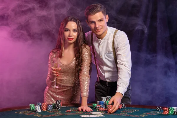 Hombre y mujer jugando al póquer en el casino, celebrando la victoria en la mesa con montones de fichas, cartas, champán. Negro, fondo de humo. Primer plano . — Foto de Stock