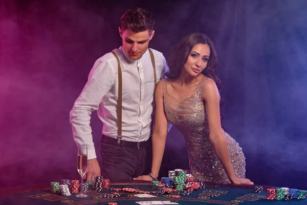 Hombre y mujer jugando al póquer en el casino, celebrando la victoria en la mesa con montones de fichas, cartas, champán. Negro, fondo de humo. Primer plano . — Foto de Stock