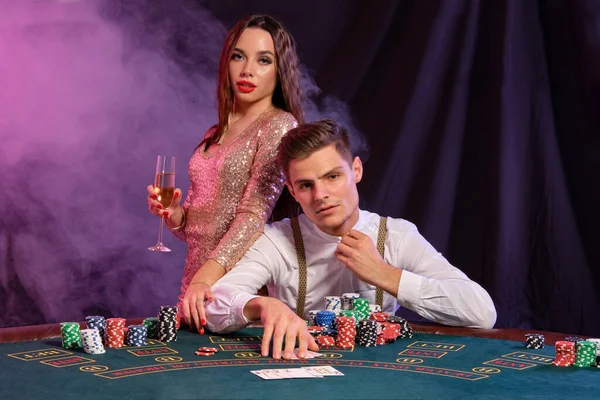 Homme jouant au poker au casino assis à table avec des piles de jetons, argent, champagne, cartes. Célébrer la victoire avec une femme. Fond noir. Gros plan . — Photo