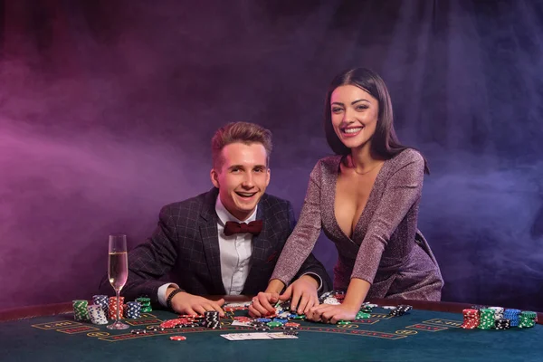 Homem jogando poker no casino sentado à mesa com pilhas de fichas, dinheiro, champanhe, cartas. A celebrar a vitória com a mulher. Fundo preto. Close-up . — Fotografia de Stock