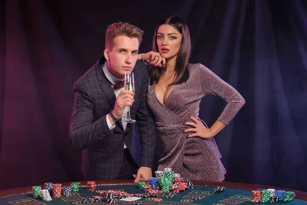 Άντρας και γυναίκα παίζουν πόκερ στο καζίνο, γιορτάζοντας τη νίκη στο τραπέζι με στοίβες από μάρκες, χρήματα, κάρτες, σαμπάνια. Μαύρο φόντο. Τζόγος. Κοντινό πλάνο. — Φωτογραφία Αρχείου