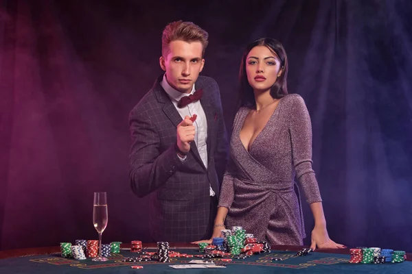 Hombre y mujer jugando al póquer en el casino, celebrando la victoria en la mesa con montones de fichas, dinero, cartas, champán. Negro, fondo de humo. Primer plano . — Foto de Stock