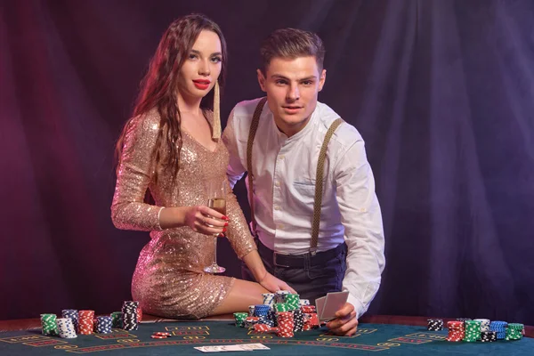 Uomo e donna che giocano a poker al casinò, celebrano la vittoria a tavola con pile di patatine fritte, carte, champagne. Nero, sfondo fumo. Primo piano . — Foto Stock