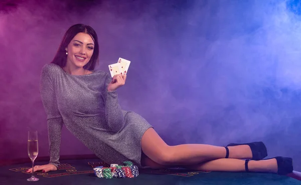 Gri elbiseli kız kumarhanede poker oynuyor, elinde kartlarla masada patates kızartması ve şampanya ile yatıyor. Siyah, dumanlı arka plan. Yakın plan.. — Stok fotoğraf