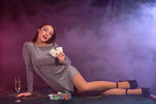 Dívka v šedých šatech hraje poker v kasinu, drží karty, leží na stole s chipsy a šampaňské na něm. Černá, kouřové pozadí. Detailní záběr. — Stock fotografie