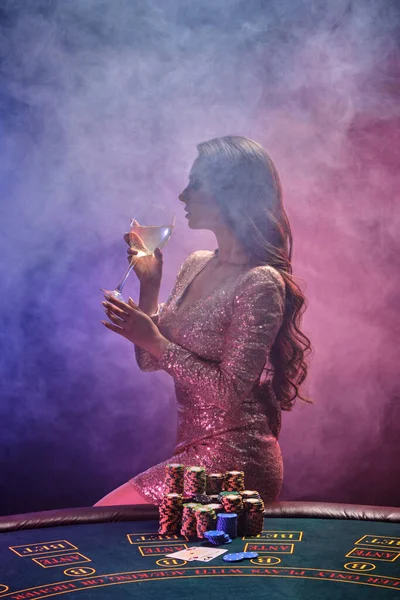 Μελαχρινή κοπέλα με τέλειο χτένισμα και φωτεινό make-up πίνει σαμπάνια σε ένα τραπέζι παιχνιδιού. Καζίνο, πόκερ. — Φωτογραφία Αρχείου