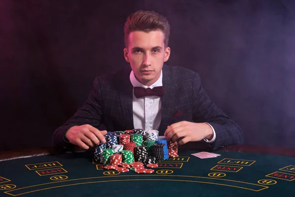Elegantní muž hraje poker v kasinu. Sedí u stolu s hromadou barevných žetonů a karet. Černá, kouřové pozadí. — Stock fotografie