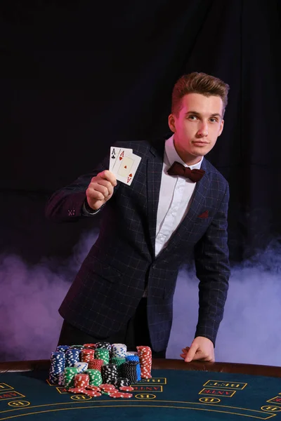 Homme élégant jouant au poker au casino. Il montre deux as tout en posant à la table avec des piles de jetons sur elle, sur fond noir . — Photo