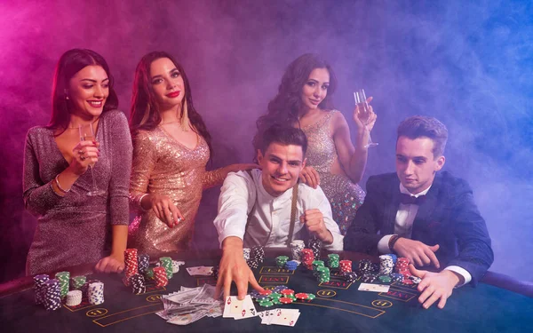 Freunde spielen Poker im Casino. Sie feiern ihren Sieg, trinken Champagner und posieren am Tisch mit einem Stapel Chips, Geld und Karten darauf. — Stockfoto