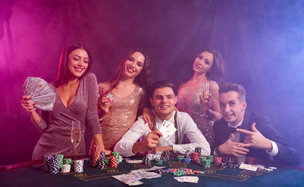 Přátelé hrajou poker v kasinu. Oslavují výhru, pijí šampaňské a pózují u stolu s hromadou žetonů, peněz a karet.. — Stock fotografie