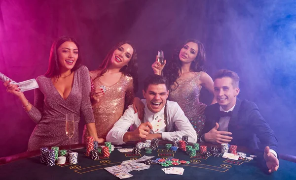 Vrienden die poker spelen in het casino. Ze vieren hun overwinning, drinken champagne en poseren aan tafel met stapels chips, geld en kaarten erop.. — Stockfoto