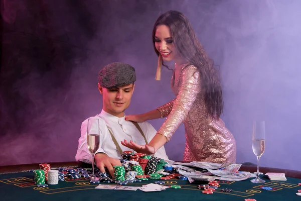 Elegant man playing poker at casino accompanied by a girl. Sitting at table with stacks of chips, money and cards on it. Black, smoke background. — Stock Photo, Image