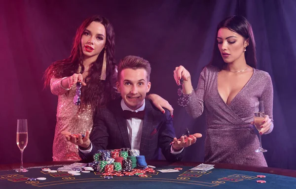 Homme élégant jouant au poker au casino accompagné de deux filles. Assis à table avec des piles de jetons et de cartes dessus. Noir, fond de fumée . — Photo