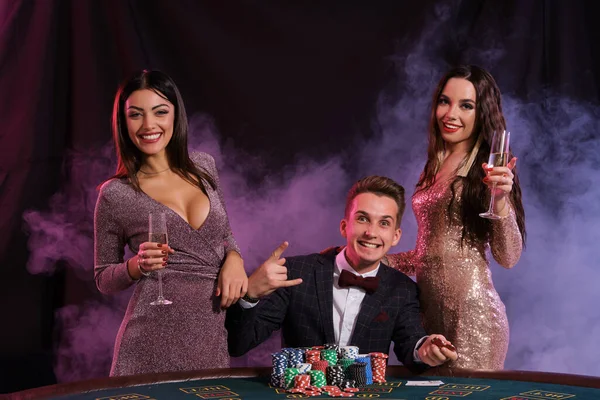 Elegant mann som spiller poker på kasino ledsaget av to jenter. Sittende ved bordet med stabler med chips og kort på. Svart, røykbakgrunn . – stockfoto
