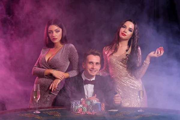 エレガントな男は2人の女の子を伴うカジノでポーカーをプレイ.テーブルにはチップやカードの山があります。黒煙の背景. — ストック写真