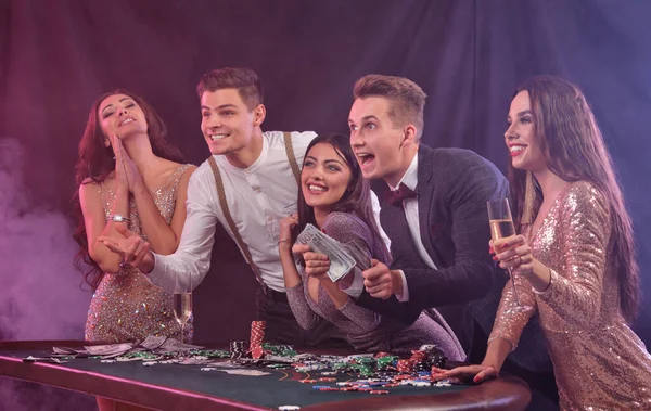 Friends playing poker at casino, at table with stacks of chips, money, cards, champagne on it. Celebrating win, excited. Black background. Close-up. — Stok fotoğraf