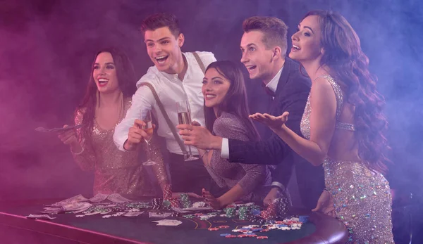 Friends playing poker at casino, at table with stacks of chips, money, cards, champagne on it. Celebrating win, excited. Black background. Close-up. — Stock fotografie
