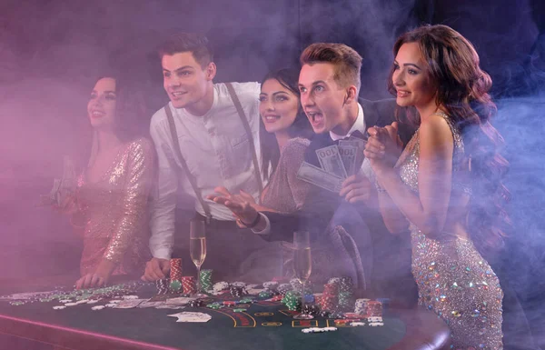 Friends playing poker at casino, at table with stacks of chips, money, cards, champagne on it. Celebrating win, excited. Black background. Close-up. — Stock fotografie