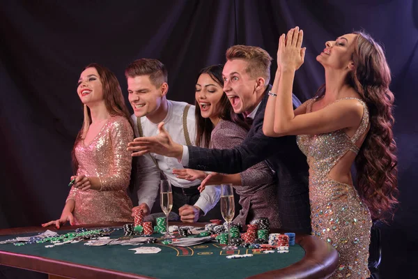 Friends playing poker at casino, at table with stacks of chips, money, cards, champagne on it. Celebrating win, excited. Black background. Close-up. — ストック写真