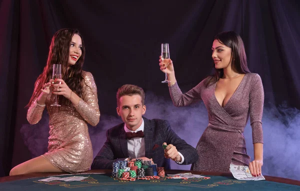 Mann, der im Casino Poker spielt, sitzt am Tisch und stapelt Chips, Geld und Karten. Mit zwei Frauen den Sieg feiern. Schwarzer, rauchiger Hintergrund. Nahaufnahme. — Stockfoto
