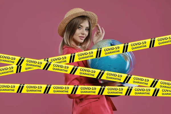 Biohazard Coronavirus yellow warning tapes against blonde woman in hat and red pantsuit. She smiling, holding a globe, posing on pink background. Worldwide pandemic COVID-19. Close up, copy space