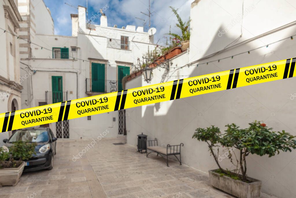 Biohazard Coronavirus warning tapes against courtyard of old town Martina Franca with parked car and houses painted in white. Apulia, Italy. Quarantine. Pandemic COVID-19, 2019-nCoV, SARS-CoV-2