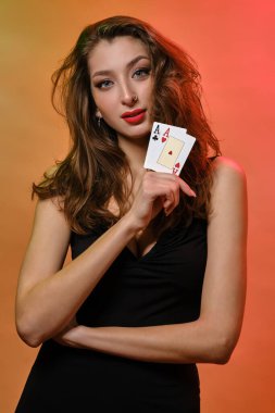 Siyah elbiseli, burnunda küpe olan esmer kız. Renkli arka planda poz veren iki as gösteriyor. Kumar, poker, kumarhane. Yakın plan.