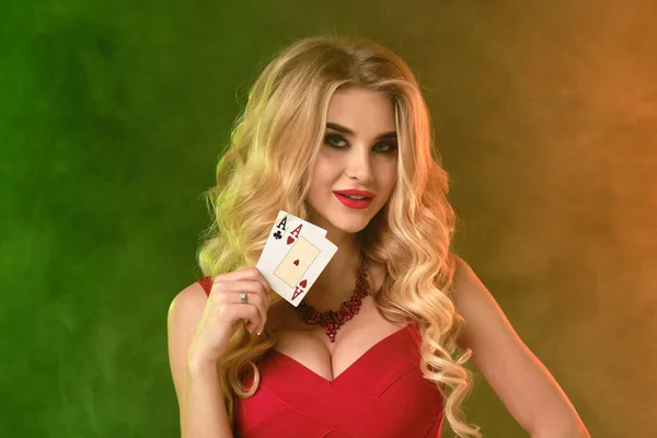 Gorgeous curly blonde model, bright make-up, in red dress and necklace. She smiling, showing two playing cards, posing on colorful smoky studio background. Poker, casino. Close-up, copy space