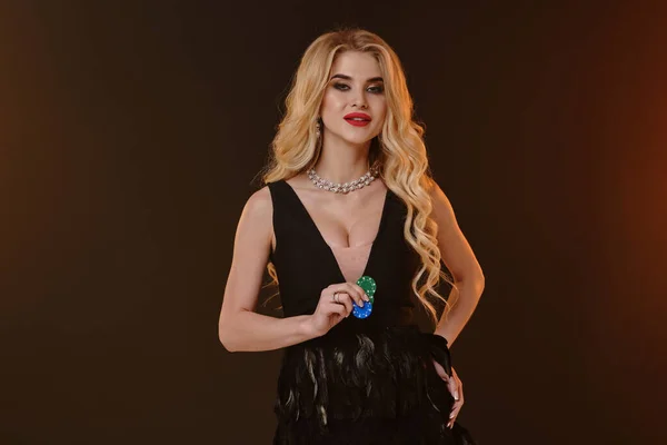 Ξανθό μοντέλο σε μαύρο κομψό φόρεμα και κολιέ. Χαμογελάει, δείχνει πράσινες και μπλε μάρκες, ποζάροντας σε πολύχρωμο φόντο. Πόκερ, καζίνο. Κοντινό πλάνο — Φωτογραφία Αρχείου
