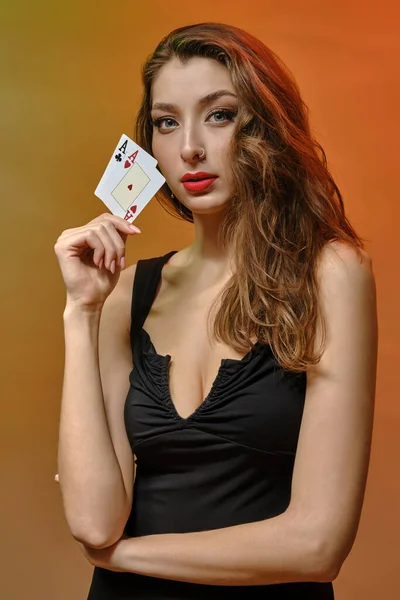 Modelka s náušnicí v nose, v černých šatech. Ukazuje dvě esa, pózuje na barevném pozadí studia. Hazard, poker, kasino. Detailní záběr — Stock fotografie