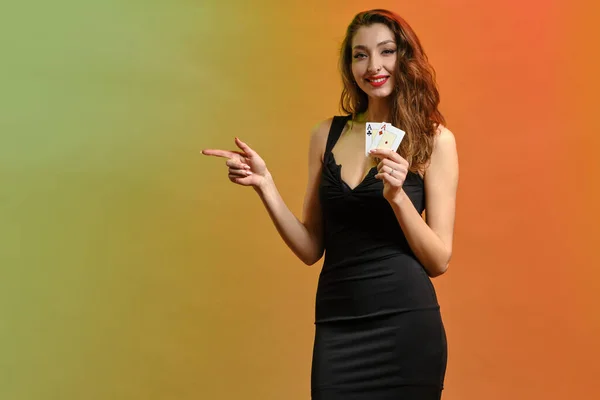 Female in black dress. Holding two aces, smiling, pointing at something, posing on colorful background. Poker, casino. Template or mockup. Close up