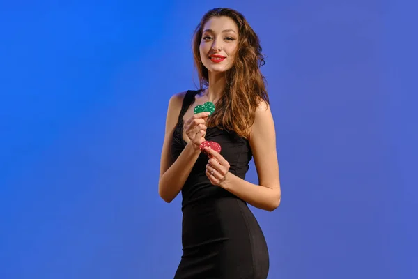Modelka s náušnicí v nose, v černých šatech. Usmívá se, drží dva zelené a červené chipsy, pózuje na modrém pozadí. Poker, kasino. Detailní záběr — Stock fotografie