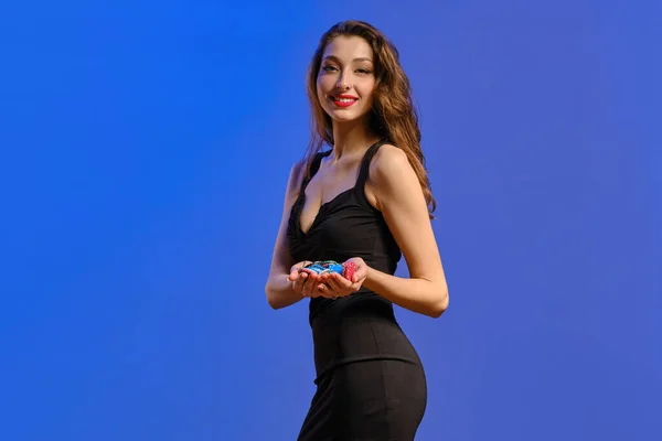 Брюнетка в черном платье. Она показывает горстку красочных чипсов, улыбаясь, позируя боком на синем фоне. Покер, казино. Крупный план — стоковое фото